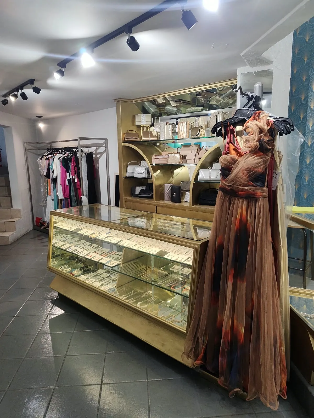 Mostrador dorado con aretes y collares, vitrina de bolsas de fiesta y un vestido de tul en maniquí